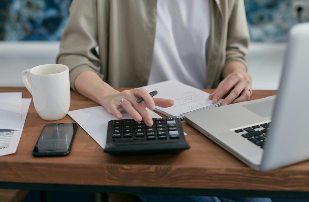 a person sitting at a wooden table using a notepad, calculator, and laptop